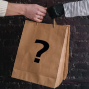 Kinsey Investigations Mystery Shopper Services - Two hands from two different people who are not pictured hold onto the handle of a brown shopping bag that is stamped with a large black question mark.