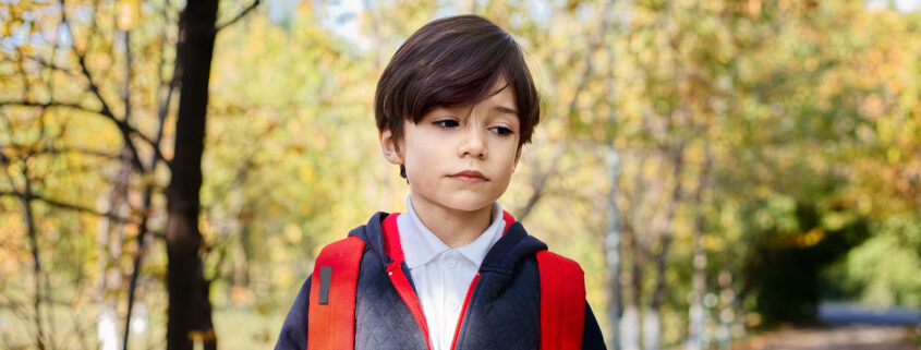 Kinsey Investigations Family Law Private Investigations Services - A school age boy is pictured walking along a tree-lined street and wearing a blue coat with a red zipper and a red backpack.