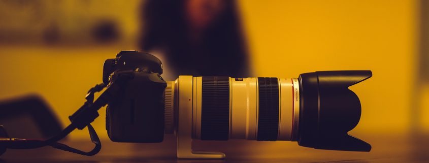 Kinsey Investigations offers Surveillance Services - A camera with a large telephoto lens is pictured in the foreground. A person with long dark hair sits in the blurred background