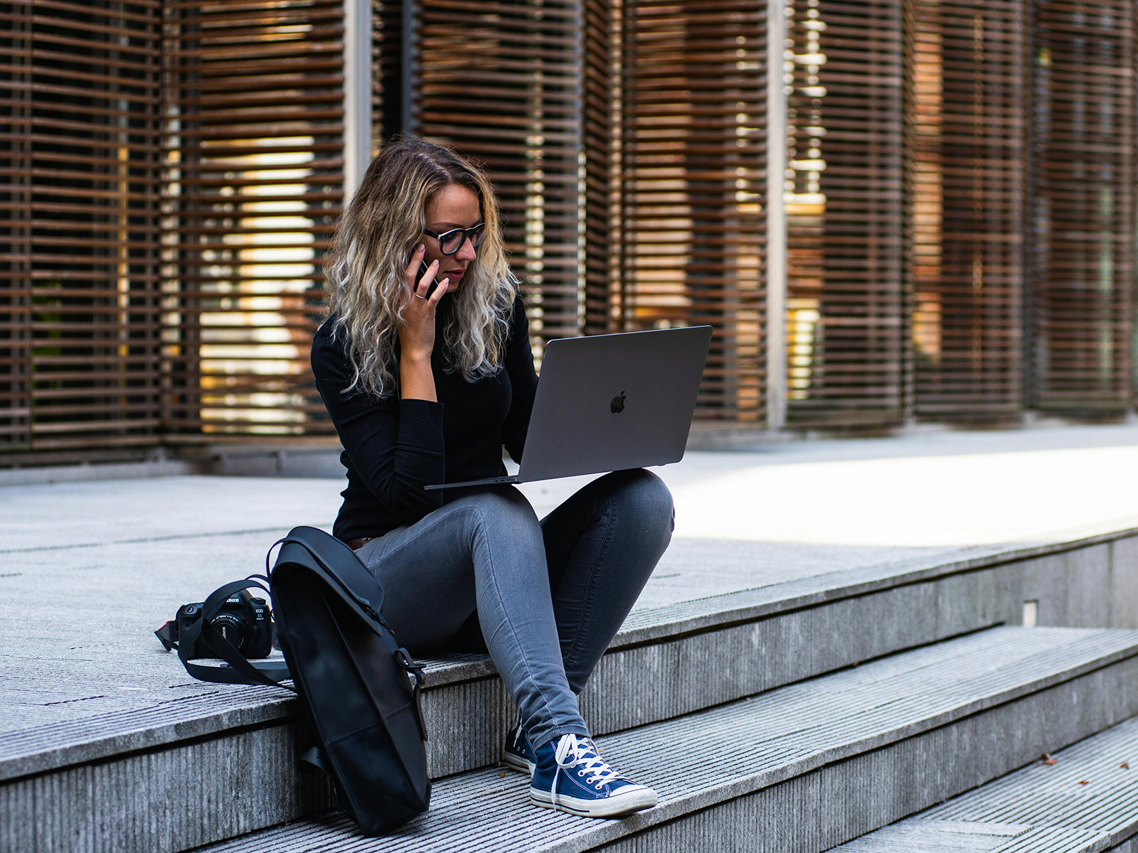KinseyInvestigations.com Becoming a Private Investigator - A woman using a cell phone and laptop sits on steps in front of a building.