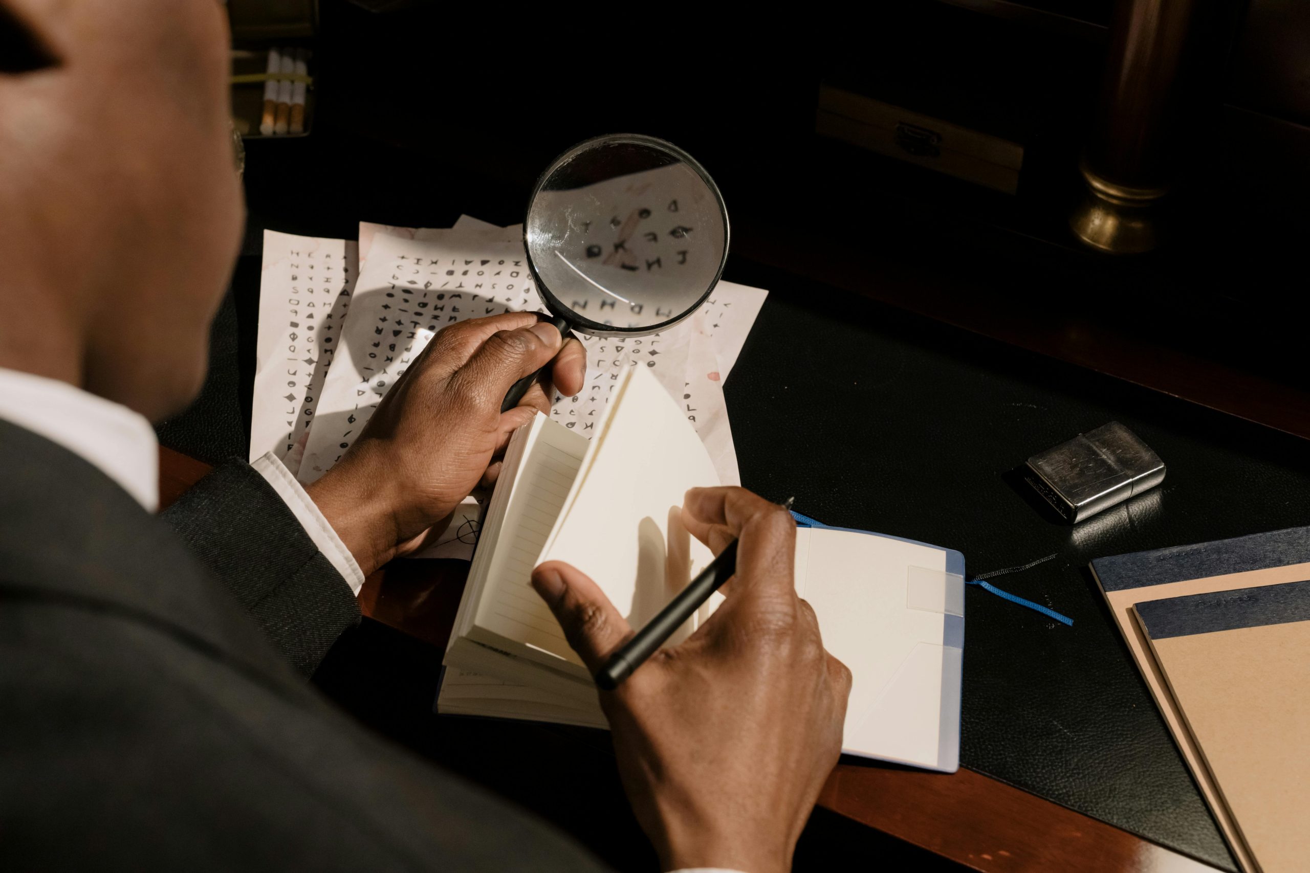 Black private detective seen from behind referencing a notebook as he attempts to decode cryptic text. He holds a magnifying glass in his left hand and a pen in his right. - Visit KinseyInvestigations.com.