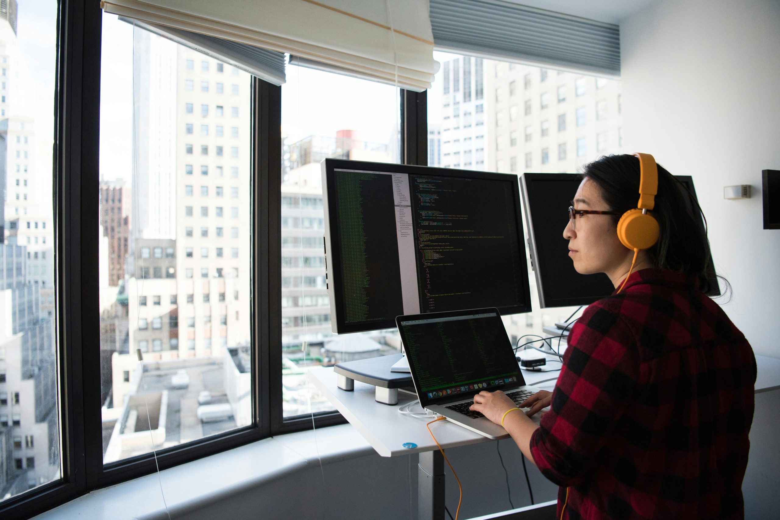 Woman standing at a computer screen display wearing yellow headphones looking out the window at the business district outside - Visit KinseyInvestigations.com.