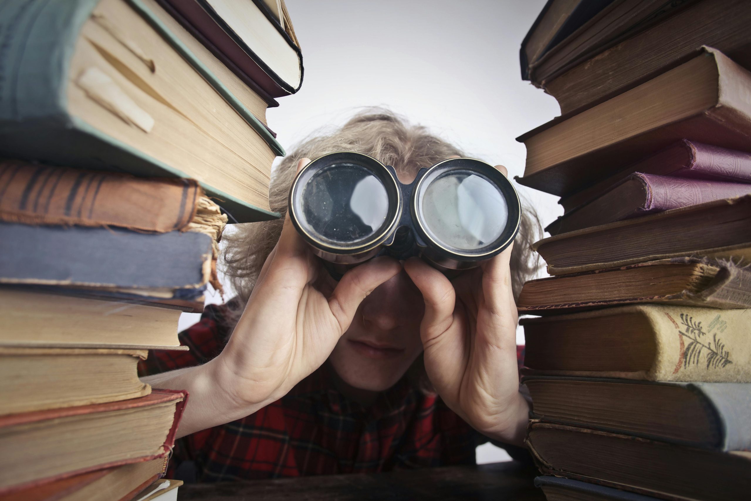 Blonde woman with binoculars peering out from between two stacks of books - Visit KinseyInvestigations.com.