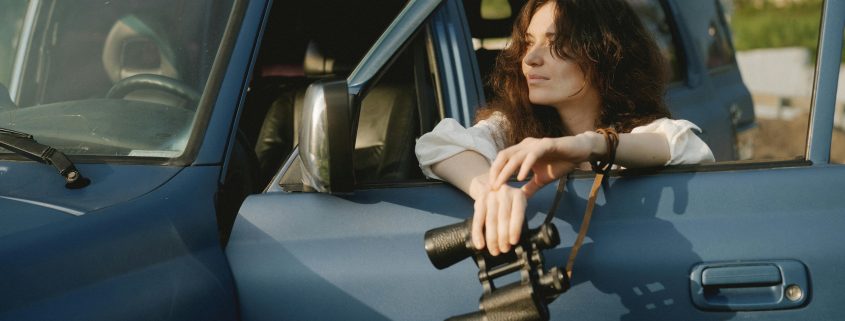 Female private detective in car window holding binoculars - Consult Kinsey Investigations.