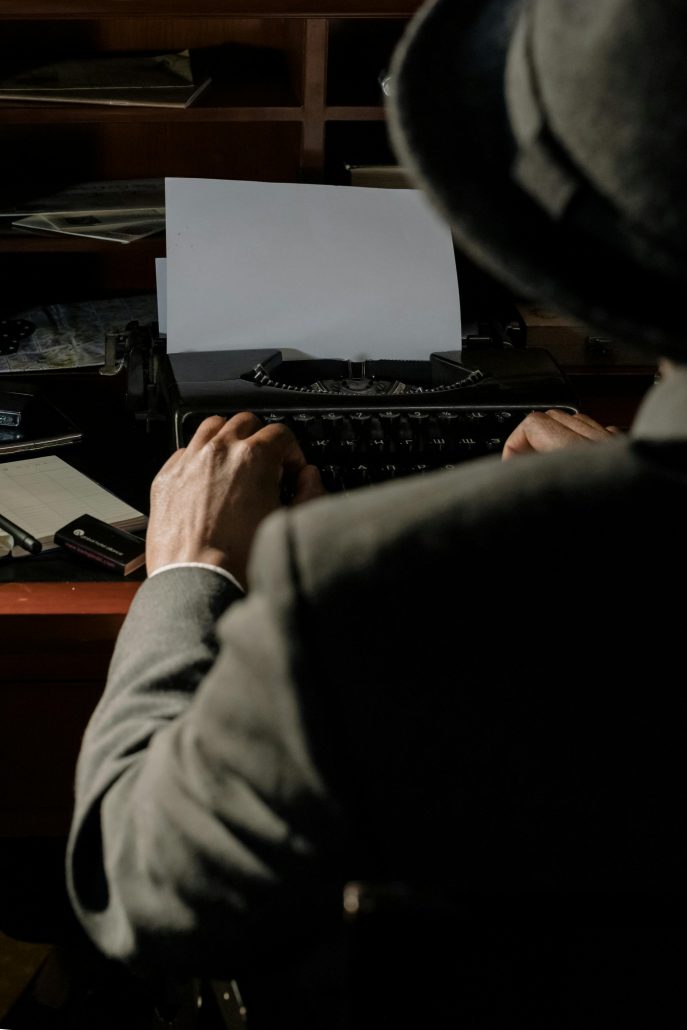 A movie private detective seen from behind over his shoulder as he types at a typewriter - Consult Kinsey Investigations.